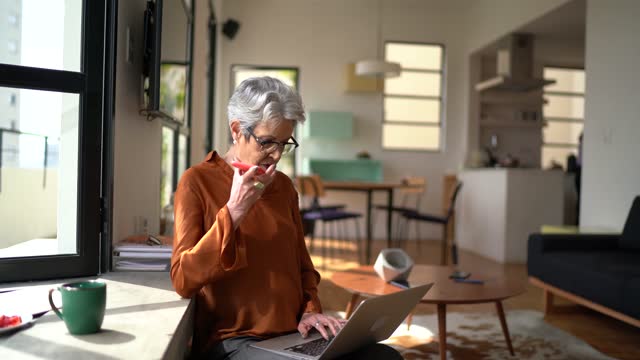 Senior woman working at home using laptop and sending audio message on smartphone