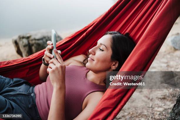 young woman texting message while relaxing on hammock at lakeshore - hängematte stock-fotos und bilder