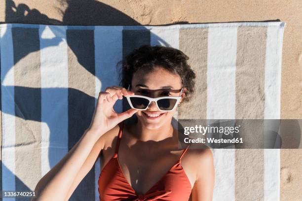 smiling young woman wearing sunglasses while lying on towel at beach - sunglasses overhead fotografías e imágenes de stock