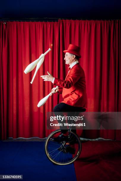 male artist juggling pins while balancing on unicycle in circus - circus performer fotografías e imágenes de stock