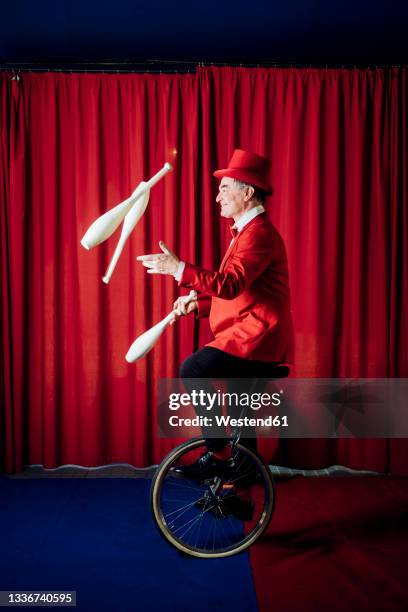 male artist juggling pins while balancing on unicycle in circus - fare il giocoliere foto e immagini stock