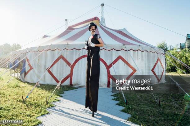 male artist in costume standing on stilts in front of circus tent - échasses photos et images de collection