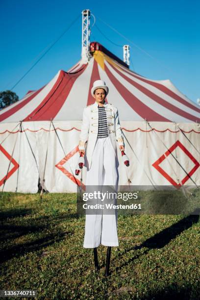 male artist holding juggling pins while standing with stilts in front of circus tent - zirkuszelt stock-fotos und bilder