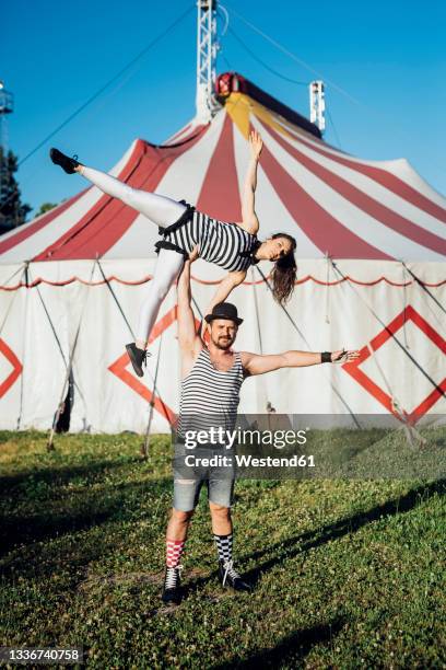 muscular male acrobat lifting female performer while standing on meadow - artista del circo foto e immagini stock