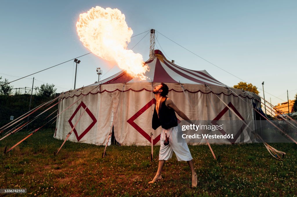 Male acrobat blowing fire while standing in front of circus tent