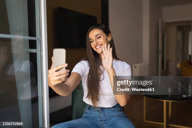 smiling young woman waving hand during video call through smart phone - brown hair waves stock pictures, royalty-free photos & images