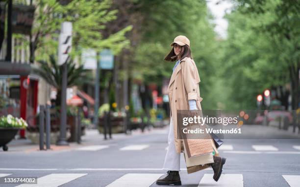 young woman with shopping bags walking on road - trenchcoat stock-fotos und bilder