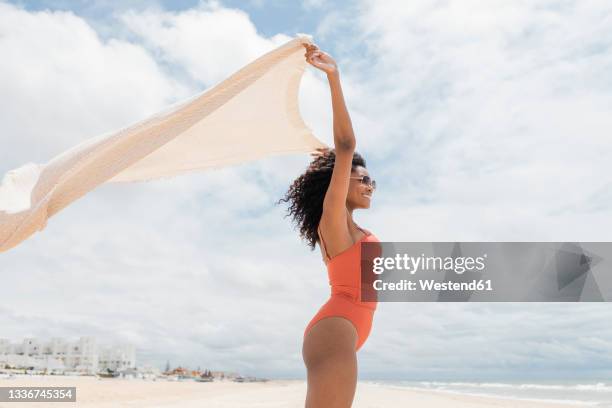 smiling woman holding towel while standing at beach - swimwear stock pictures, royalty-free photos & images