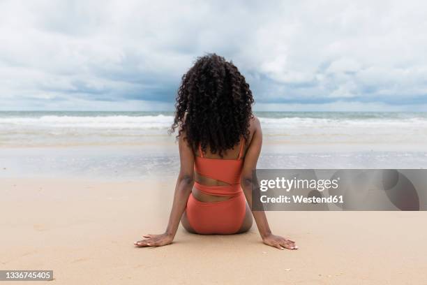 young woman looking at sea while sitting on sand - black hair imagens e fotografias de stock