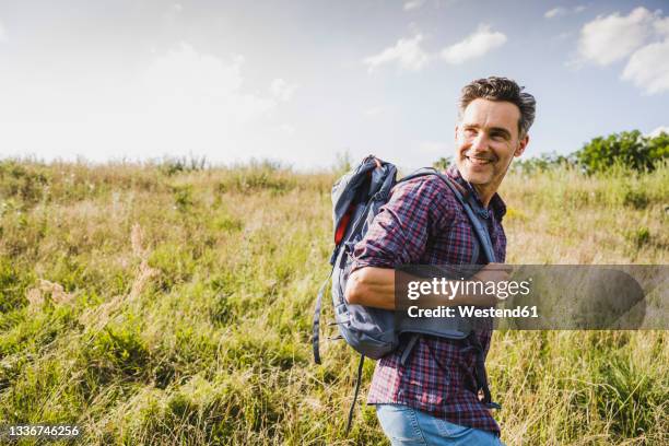 smiling man with backpack hiking at meadow on sunny day - man looking over shoulder stock pictures, royalty-free photos & images