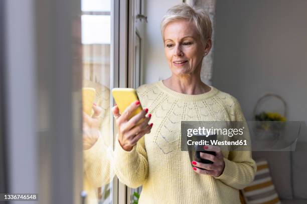 mature woman using smart phone while holding mug at home - middle aged woman at home stock pictures, royalty-free photos & images