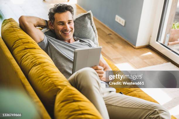 smiling man using digital tablet while lying on sofa at home - man couch foto e immagini stock