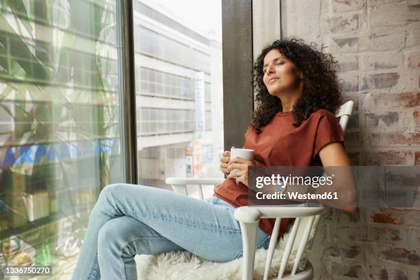 relaxed businesswoman holding coffee cup while sitting with eyes closed on chair by window - coffee break office ストックフォトと画像