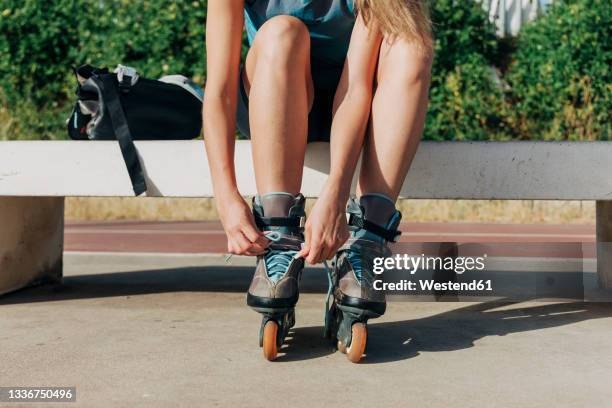 young woman tying shoelace of inline skate while sitting on bench during sunny day - inline skating fotografías e imágenes de stock