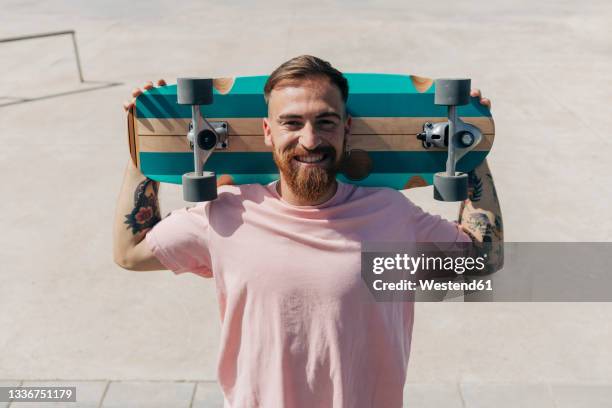 happy bearded young man carrying skateboard on shoulders - skatepark imagens e fotografias de stock