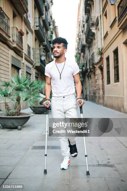 young man walking in alley with help of crutches amidst buildings - latina legs fotografías e imágenes de stock