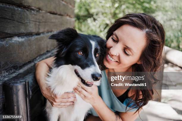 smiling woman looking at dog while hugging by cottage in forest - border collie stock pictures, royalty-free photos & images