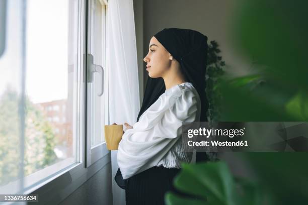 young female freelancer having coffee while looking through window at home - arabische frau kopftuch stock-fotos und bilder