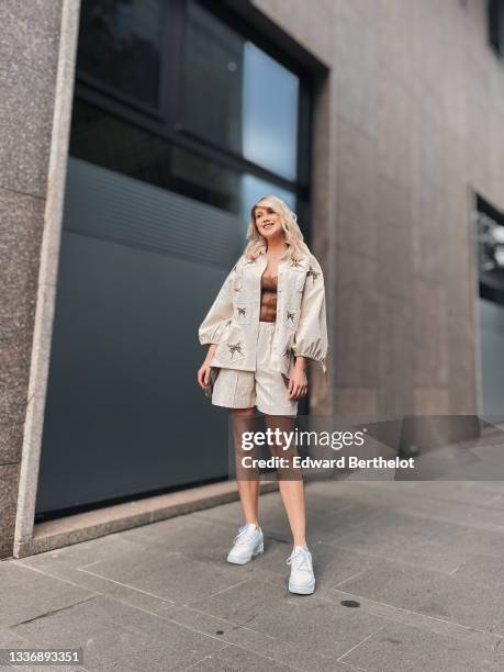 Andra Morariu wears a silver chain with cross pendant necklace, a brown shiny leather shoulder-off corset t-shirt, a beige shiny with brown...