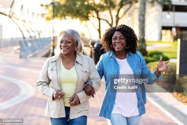 two senior african-american women walking arm in arm - two elderly friends chatting in a community park stock pictures, royalty-free photos & images