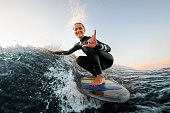 smiling woman sits on wakesurf board and rides the wave and touches the waves with one hand
