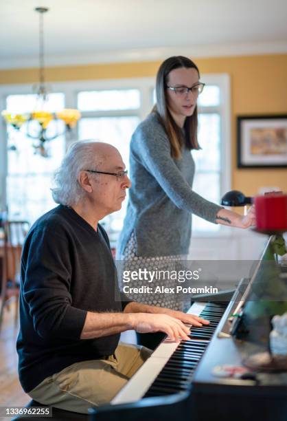 hombre mayor de cabello blanco de 72 años, padre, tocando el piano, y su hija adulta lo ayudan pasando las páginas de la partitura. - 26 years old fotografías e imágenes de stock