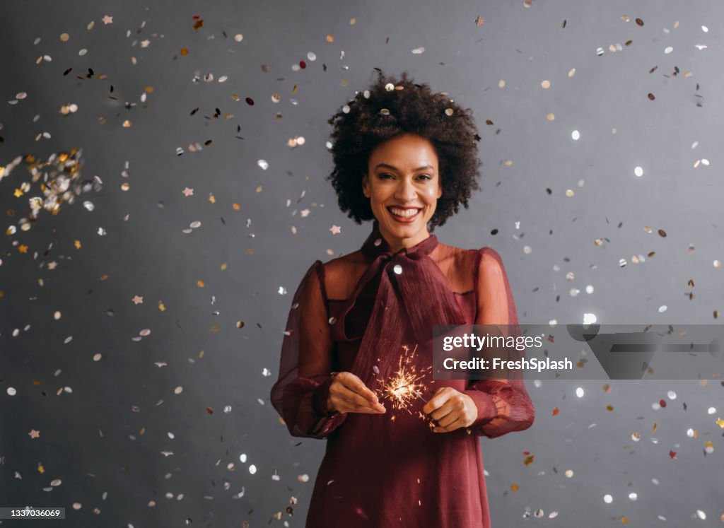 It's Time to Celebrate: Portrait of a Beautiful Afro American Woman Holding a New Year's Sprinkler in Confetti Rain (Gray Background)