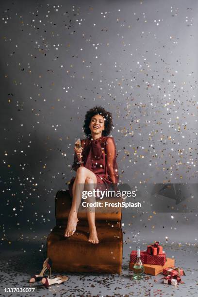 new year's celebration: beautiful afro american woman sitting barefoot with her legs crossed, holding a glass of champagne in confetti rain - new year gifts imagens e fotografias de stock