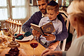 Small Jewish boy and his father reading Tanakh at dining table during Hanukkah.