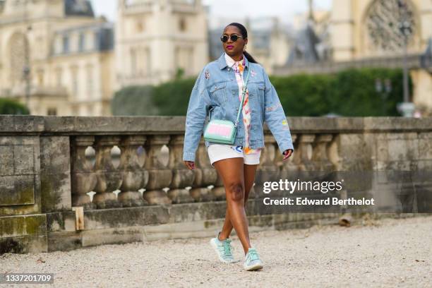 Jennifer Joseph wears black sunglasses, gold large pendant earrings, a white with multicolored paint pattern shirt, a blue faded denim oversized...