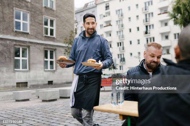 food truck owner bringing burgers to customers sitting at tables - food truck street stock-fotos und bilder