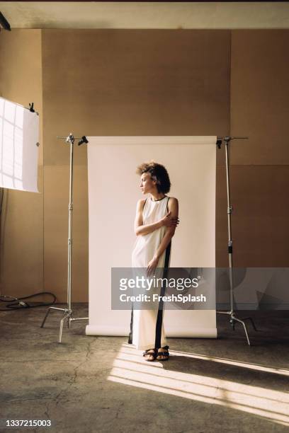 vertical shot of a beautiful mixed race mid adult woman standing in front of a white backdrop in a photo studio, looking away - beige dress stock pictures, royalty-free photos & images