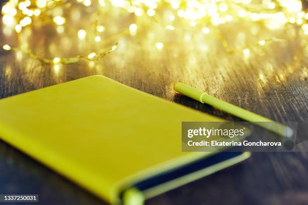 a yellow closed notebook on a wooden table with a yellow pen against a background of sparkling christmas lights, golden glitter and garlands. - wensen stockfoto's en -beelden