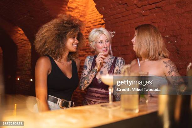 smiling diverse girlfriends with tattoos talking at bar counter - cocktail counter stock pictures, royalty-free photos & images