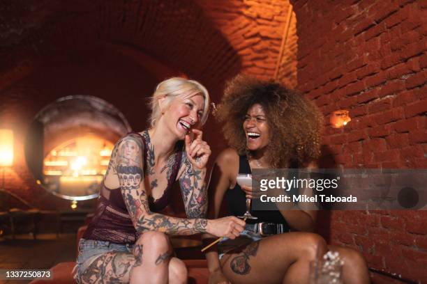 happy tattooed multiracial girlfriends with cocktail talking in pub - lesbe stock-fotos und bilder