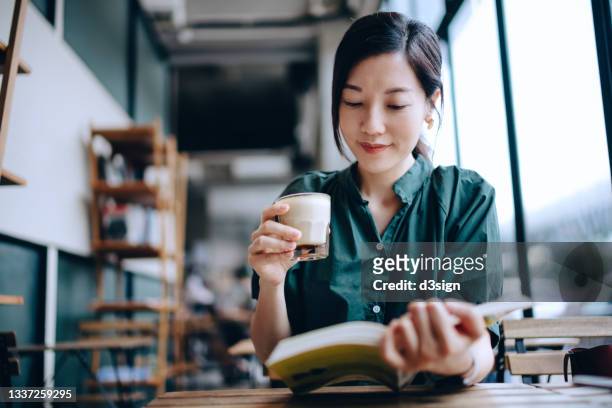 young asian woman taking a break, having a quiet time enjoying a cup of coffee and reading book in cafe - chino oriental fotografías e imágenes de stock
