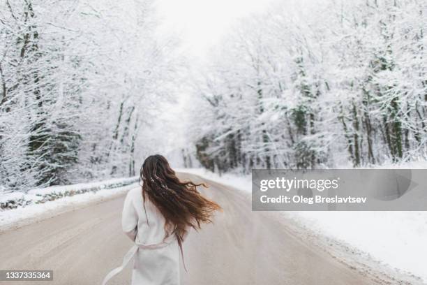 beautiful happy woman walking by snowy winter road in forest. view from behind, unrecognisable person - snowy back alley stock pictures, royalty-free photos & images