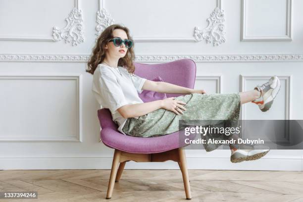 young fashionable woman model posing sitting on an armchair in the studio in trendy modern clothes from the new collection catalog - elegant fashion stockfoto's en -beelden