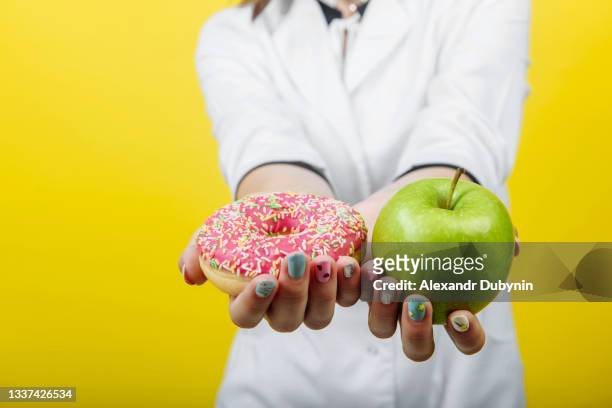 doctor nutritionist holding a sugar donut and a green apple on a yellow background. the concept of diet comparison. free place - natuurlijke staat stockfoto's en -beelden