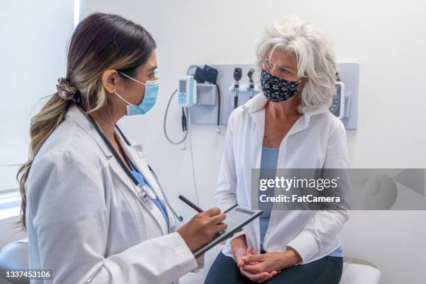 masked senior woman listening to her doctor during a check up appointment - fatcamera doctor stock pictures, royalty-free photos & images