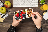 First person top view photo of woman's hands holding lunchbox with healthy meal nuts and berries over apples glass of juice flowerpot stationery keyboard mouse on isolated dark wooden table background