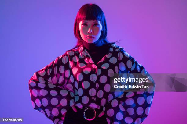 neon blue purple lights posing in studio asian chinese female fashion model with polka dot blouse in studio portrait arms akimbo - high fashion model stock pictures, royalty-free photos & images