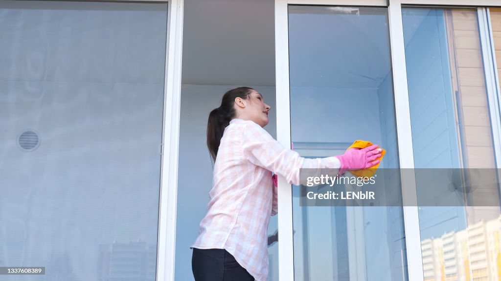 Brunette woman in gloves with spray and a rag washes the window, outside view