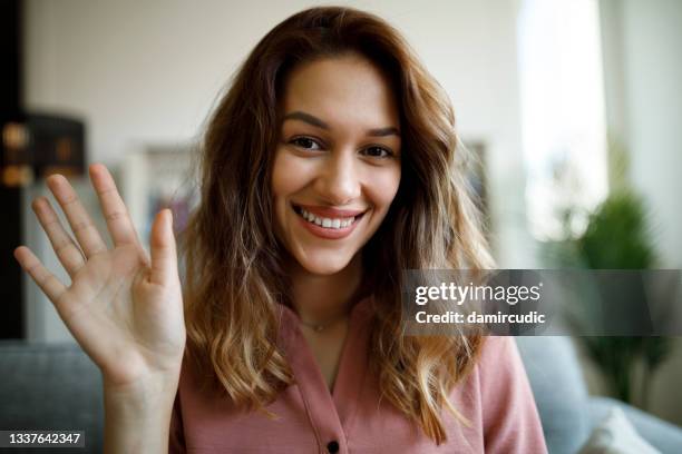 joven mujer sonriente saludando con la mano en una videollamada en la oficina en casa - businesswoman couch fotografías e imágenes de stock