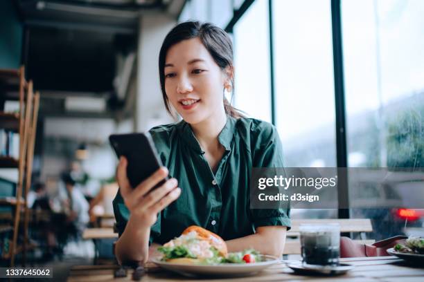 beautiful young asian woman managing online banking with mobile bank apps on smartphone while sitting in cafe having lunch. checking financial data, transferring money, paying bills and checking balances. smart banking with technology - coffee shop chat stock-fotos und bilder