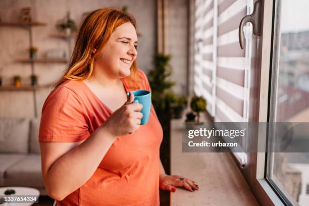 la mujer mira por la ventana y toma café - gordo fotografías e imágenes de stock