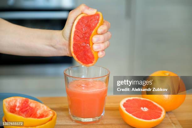 a girl or woman squeezes the juice from a red ripe grapefruit, squeezes the fruit with her hand. a glass on a cutting board, against the background of a wooden kitchen table. i'm making juice for breakfast. the concept of vegetarian, vegan and raw food. - immune system stock pictures, royalty-free photos & images