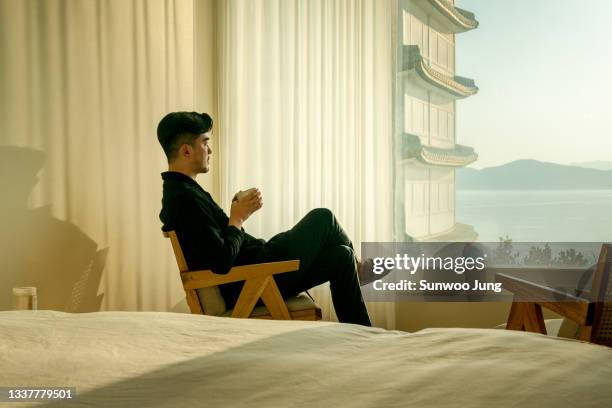man with mug looking outside in hotel room - asiatische kultur stock-fotos und bilder