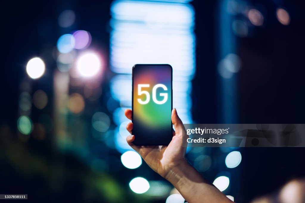 Close up of woman's hand using smartphone against illuminated financial district in the city, with the concept of 5G communications technology