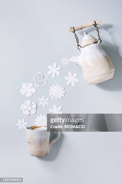 Creative layout with variety of white paper flowers flowing from ceramic teapot to cup over gray background. Flat lay. Copy space. Flower tea. Summer...